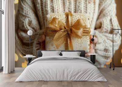 A woman in a voluminous sweater, Christmas atmosphere vibe against of festive background, bokeh garland lights. Hands holding a beige craft present box with a golden ribbon and bow, warm colors Wall mural