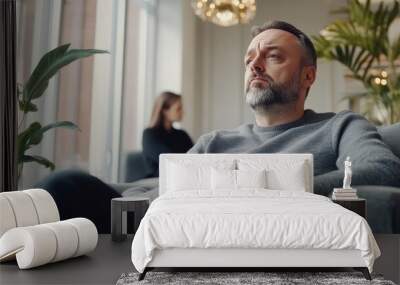 A contemplative man with light beard, lounging comfortably on a gray sofa in a modern room with large windows and natural light, embodying introspection and calmness. Wall mural