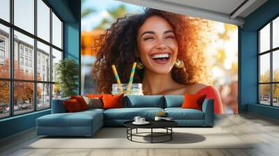 A laughing woman with exuberant curly hair holds a green smoothie, enjoying a sunny day outdoors, exemplifying joy, health, and vibrant energy of a lively lifestyle. Wall mural