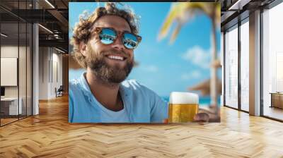 A man with curly hair and sunglasses enjoys a refreshing cold drink, possibly beer, at a sunny resort beach, smiling under the clear blue sky and palm trees. Wall mural