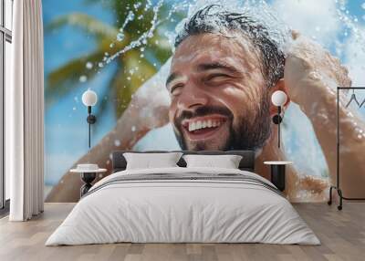 A smiling man enjoys a refreshing outdoor shower under the clear sky, with water streaming down his face and palm trees in the background, depicting a lively summer moment. Wall mural