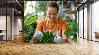 A woman wearing an orange shirt and apron carefully tends to vibrant green plants in a lush indoor setting, showcasing a passion for gardening and nature. Wall mural