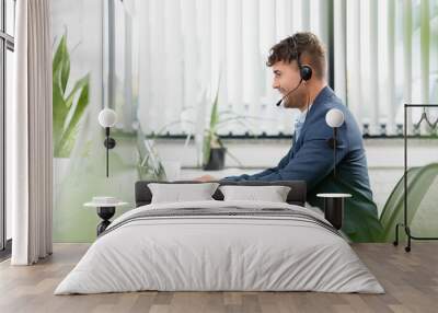  man in headset typing on laptop, while sitting at workplace with plants in office on blurred foreground Wall mural