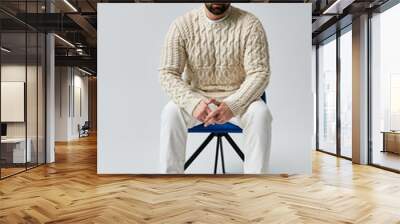 A bearded man in a white sweater striking a pose while sitting on a blue chair in a studio with a grey background. Wall mural