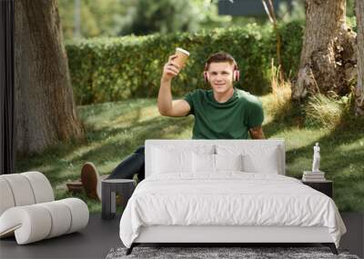 A cheerful young man with a prosthetic leg sits on the grass, sipping a drink and enjoying the outdoors. Wall mural