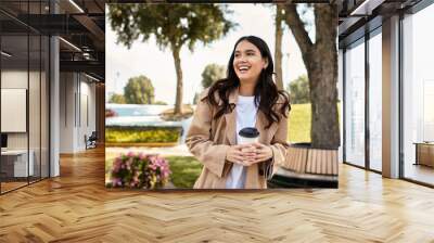 A cheerful young woman dressed in cozy autumn attire smiles while sipping her warm drink outdoors. Wall mural