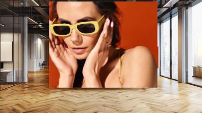 A fashionable woman wearing yellow sunglasses, hands on face, in studio on orange background. Wall mural