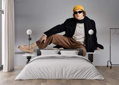 A fashionable young man sits elegantly in a black coat and yellow beanie, showcasing his unique style. Wall mural