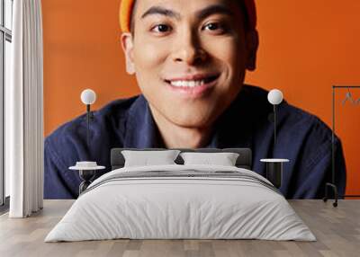 A handsome Asian man exudes style in a blue shirt and orange hat against a vibrant orange backdrop. Wall mural