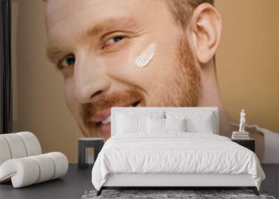 A handsome man in casual attire happily wears a facial cream for skincare. Wall mural