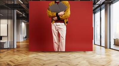 A handsome young man stands poised, cradling two laptops in his arms, dressed in a trendy yellow jacket. Wall mural