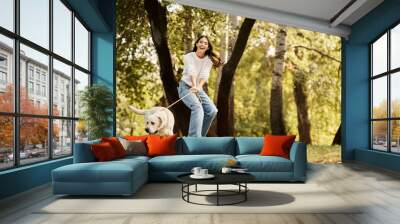 A joyful young woman in cozy autumn attire plays with her dog amidst fall colors. Wall mural