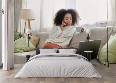 A joyful young woman relaxes at home, sipping tea while chatting on her phone and using her laptop. Wall mural