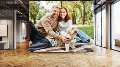 A loving couple and their dog sit on a checkered blanket in a park. Wall mural