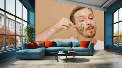 A man in casual attire gently using face roller for a skincare routine. Wall mural