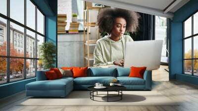 A multicultural woman engrossed in work, seated in front of her laptop computer in a modern coworking space. Wall mural