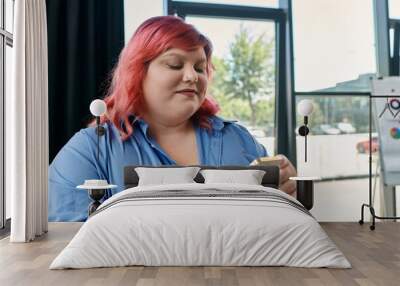 A plus size woman with pink hair and a blue shirt checks her phone in front of a window. Wall mural