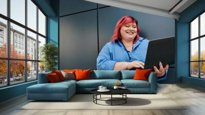 A plus size woman with pink hair smiles as she uses a laptop in front of a gray, modern wall. Wall mural