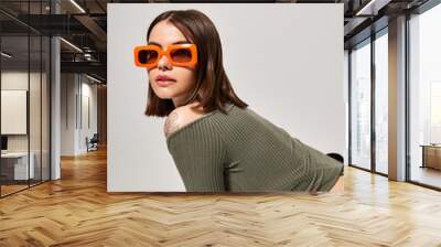 A stylish brunette woman standing confidently in a green top and trendy orange sunglasses. Wall mural