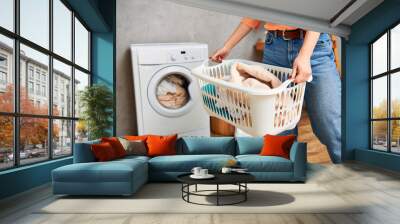 A stylish woman in casual attire holds a laundry basket in front of a washing machine. Wall mural