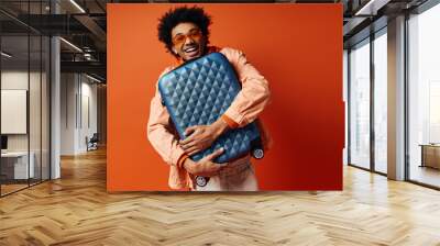 A stylish young African American man with curly hair and trendy attire stands against an orange background, holding a blue piece of luggage. Wall mural