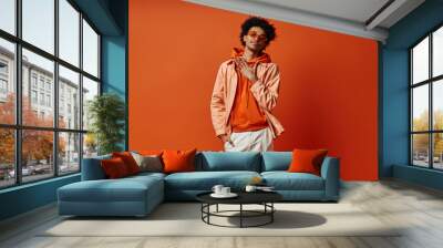 A trendy, curly African American man poses in an orange jacket and white pants against a bold orange backdrop, exuding confidence and style. Wall mural