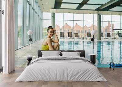 A young, beautiful brunette woman in a swimsuit sits gracefully on the edge of an indoor spa swimming pool, basking in relaxation. Wall mural