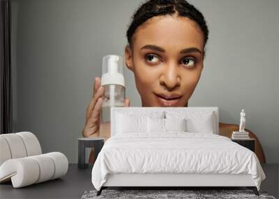 A young African American woman in a strapless top holds a bottle of liquid in front of her face on a grey background. Wall mural