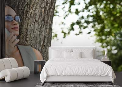 A young brunette woman with short stature stands under a tree, smiling while engaged in an outdoor phone conversation on a sunny day. Wall mural