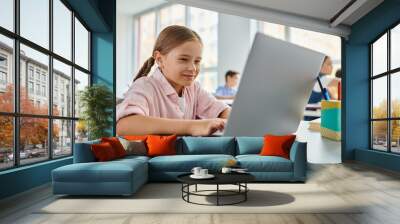 A young girl sits at a table, focused on her laptop computer Wall mural