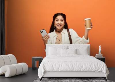 A young indian woman holds a cup of coffee and a phone against an orange background. Wall mural