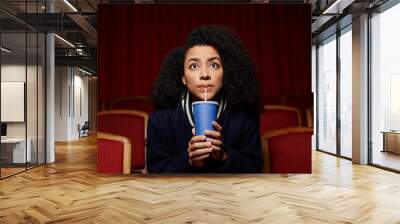 A young woman in casual clothes sits in a theater seat, holding a cup and staring up in surprise, seemingly engrossed in a movie. Wall mural