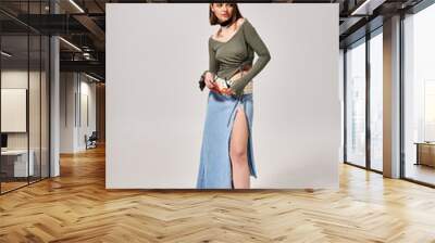 A young woman with brunette hair poses stylishly in a skirt and boots in a studio setting. Wall mural