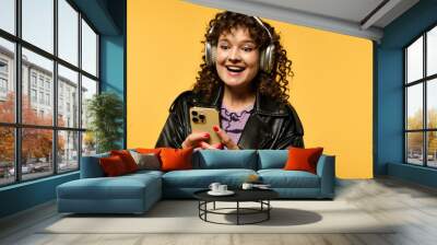 A young woman with curly hair smiles while wearing headphones and a black leather jacket against a yellow background. Wall mural