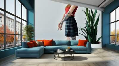 A young woman with tattoos on her arms and legs poses against a grey wall, wearing a plaid skirt and a red tank top. Wall mural