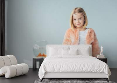 adorable kid in faux fur coat and skirt looking at camera and standing near to stool with fishbowl isolated on grey Wall mural