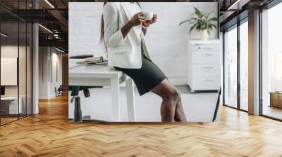 african american adult businesswoman in white formal wear leaning on office desk, smiling and holding cup of coffee Wall mural