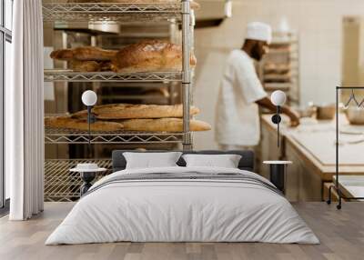 african american baker preparing raw dough at workplace with shelves of fresh loaves of bread on foreground Wall mural