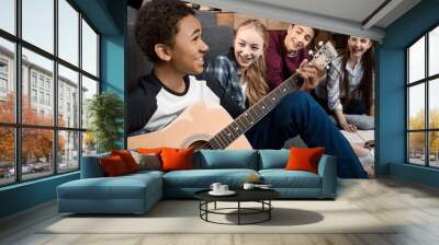 african american boy playing acustic guitar and singing while his friends listening at home, teenagers playing guitar concept Wall mural