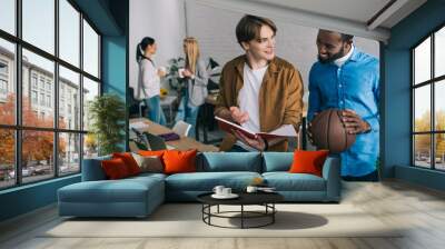 african american businessman holding ball and talking to partner with textbook and two businesswomen standing behind with coffee Wall mural