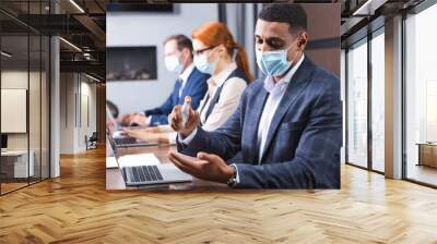 African american businessman in medical mask applying sanitizer on hands at workplace on blurred background. Wall mural