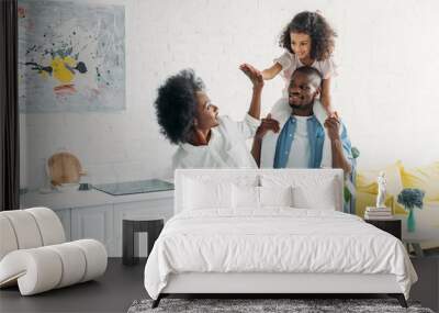 african american daughter sitting on fathers shoulders and giving high five to mother in kitchen at home Wall mural