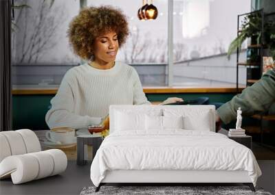 An African American woman sits at a table, enjoying a plate of food in a modern café setting. Wall mural