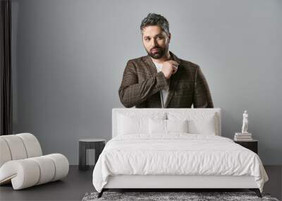 An alluring man with a beard strikes a pose in elegant attire against a grey studio background. Wall mural