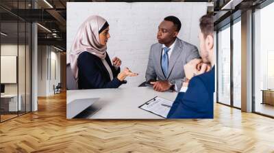 arabian businesswoman pointing with hand during meeting with multicultural business partners near contract, blurred foreground Wall mural