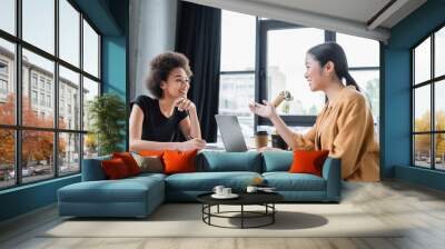 asian businesswoman talking to smiling african american colleague near laptop with blank screen Wall mural