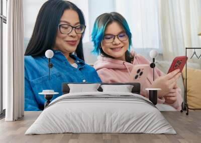 Asian mother and teenage daughter, in casual wear, sitting on a couch, focused on a cell phone screen in a cozy home setting. Wall mural