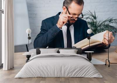 bearded lawyer in suit touching glasses while reading book in office Wall mural