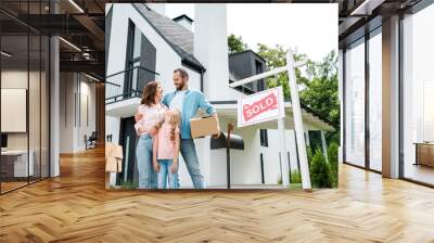 bearded man holding box and standing with wife and daughter near house and board with sold letters Wall mural