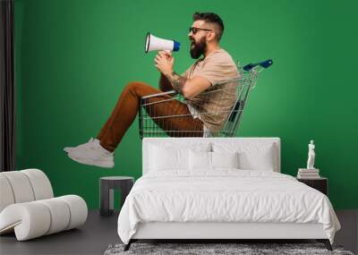 bearded man shouting into megaphone while sitting in shopping cart isolated on green Wall mural